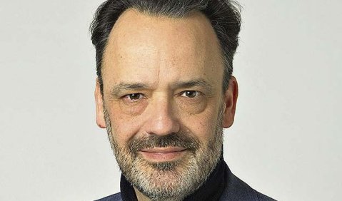 Ein Mann mittleren Alters, mit leicht grauem Bart, sonst kurzen schwarzen Haaren. Er trägt ein blaues Sakko und einen Pullover mit kurzem Rollkragen. Er lächelt leicht.