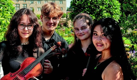 Wahrscheinlich ein junges Streichquartett oder Klavierquartett: Vier junge Musiker*innen, eine mit einer Note, eine mit einer Geige. Im Hintergrund ein Altgebäude, ähnlich einer Schule aus den späten 19. Jahrhundert