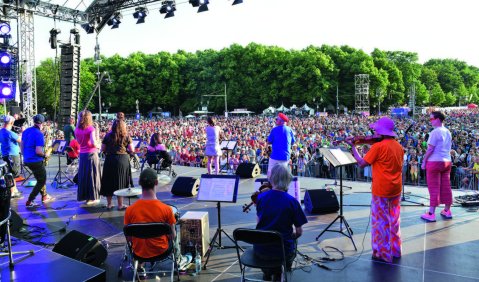 Eine Band in bunten Klamotten, dahinter das sehr sehr große Publikum des Open-Air Konzerts