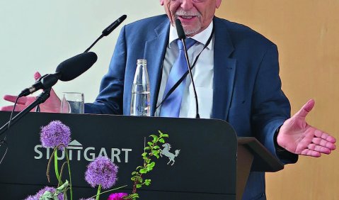 Ein älterer Mann hinter einem modernen Rednerpult. Er trägt einen blauen Anzug und sein weißes Haar als zurückhaltende Halbglatze.