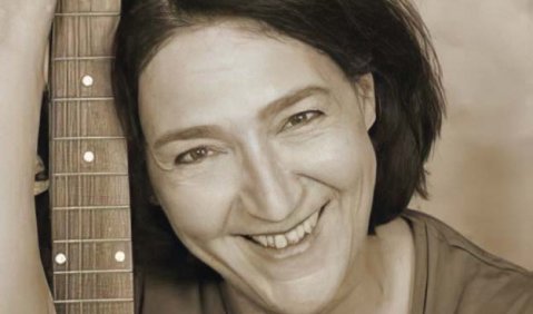 Eine lächelnde Frau lehnt auf einem Schwarz-Weiß-Foto mit Braunstich ("Sepia"). Sie hat dunkle halblange Haare und lächelt breit.