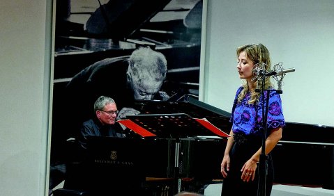Eine junge Sängerin mit langen blonden Haaren und ein älterer Pianist mit grauen Haaren musizieren zusammen.