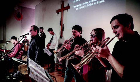 Ein Band mit Bläsern, Sänger und Keyboard performt vor einem Kreuz und einer Wand an Sätze angestrahlt sind.