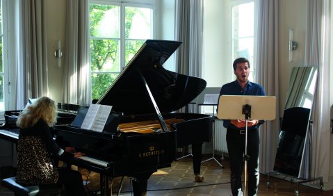 Ein Liedduo beim Musizieren. Im Hintergrund ein Fenster mit Blick ins Grüne.