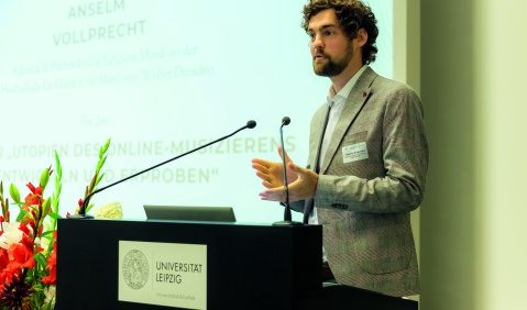 Ein junger Mann mit dunklen Locken und Bart in einem hellen Sakko vor einer Leinwand aber hinter einem Rednerpult.