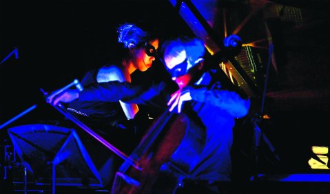 Ein Cello-Klavier-Duo im Dunkeln, nur mti blauem und etwas rotem Licht. Beide tragen schwarze Masken, die venezianischen Masken nachempfunden sind.