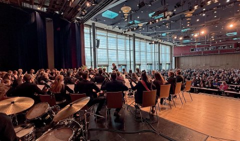 Ein großer Saal schräg hinterm Orchester ins Publikum hinein fotografiert. In der Ecke unten links sieht man ein Drumset.