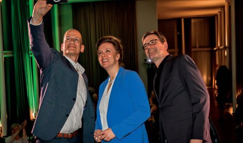 Die in der Bildbeschreibung erwähnten Personen machen ein Selfie zusammen. Alle drei tragen Anzug.