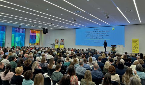 Ein mittelgroßer Vortragssaal: Voll besetzt, weiße Wände und Decke mit einer breiten Leinwand für die Präsentation.