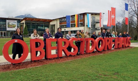 Mehrere Menschen hinter etwa ein-Meter-großen roten Buchstaben: "OBERSTDORFHAUS". Im Hintergrund sieht man ein öffentllichen Neubau mit diversen National- und Regionalflaggen davor.
