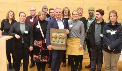 Ein Dutzend in gedeckten Farben gekleidete Menschen mit einem Mann mittleren Alters in der Mitte. Der Mann hält den Preis (eine gegossene Tafel mit 3 Bäumen im Profil).