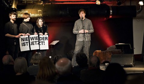 Ein Mann in grau steht im Jazzklub hinter dem Mikrofon. Neben ihm drei in schwarz gekleidete Menschen die jeweils kleine Transparente halten die zusammen den Schriftzug "NIE WIEDER IST JETZT!" ergeben.