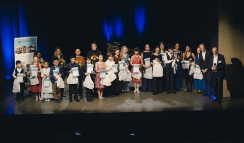 Eine große Gruppe Kinder, Jugendlicher, junger Erwachsener und Erwachsener steht auf einer Bühne. Die Menschen auf der Bühne sind schick angezogen und äußerlich von verschiedensten Ethnien.