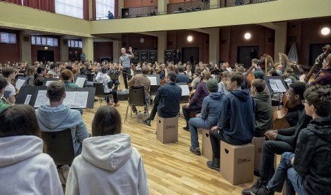 Eine hohe Aula oder ein Foyer. Schüler:innen auf Hockern und mitten zwischen Ihnen das BJO.