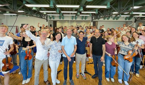Das Gruppenbild von Orchestermitglieder:innen in Alltagskleidung mehrheitlich mit Instrumenten