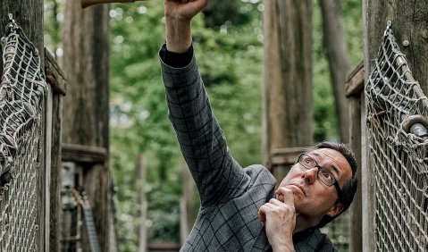 Ein Mann in grauem Anzug der in einer wundersam gezimmerten Umgebung im Wald unter freiem Himmel sitzt. Der Mann im mittleren Alter reckt mit einer Hand eine Blockflöte in die Luft. Er schaut zu ihr. Im Hintergrund das Grün der Bäume.