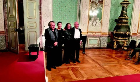 Die Vier stehen in einem barock anmutenden Saal mit feinem Parkett, rotem Teppich, fein verziertem Türrahmen und einem aufwändigen kegelförmigen Ofen.