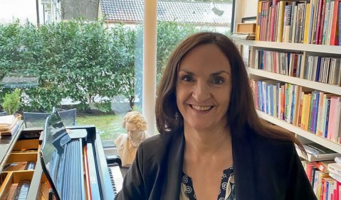 Die Frau mittleren Alters mit langen braunen Haaren lehnt entspannt lächelnd an einem Flügel.