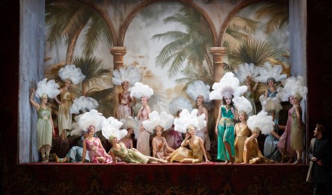 Eine Gruppe von jungen Frauen in langen Kleidern und Kopfbedeckung aus wei&szlig;en Federn formiert sich vor einem Hintergrund aus Palmen und S&auml;ulen. &copy; Monika Rittershaus