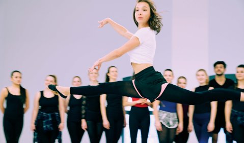 Die Balletttänzerin springt mit einem Spagat im Vordergrund. Oberkörper und Arme sind von der Beinbewegung scheinbar unberührt. Im Hintergrund schaut die restliche Ballettklasse zu.