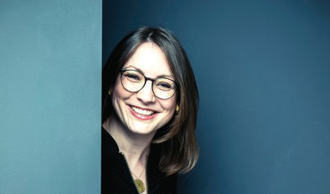 Die Frau niedrigen mittleren Alters schaut lächelnd mit etwas über schulterlangen, schwarzen Haaren und Brille zwischen zwei blauen Wänden hervor.
