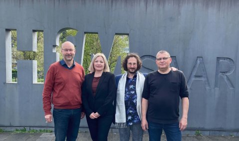 Die vier Menschen stehen draußen vor einer Betonmauer in die der Schriftzug der Hochschule eingefräst ist.