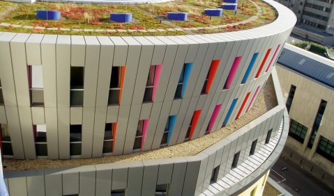 Ein breites Gebäude, ähnlich einem niedrigen runden Turm mit einem Loch/Hof in der Mitte. Die verschiedenen Stockwerke sind teils auch Achteckig oder als breiterer Kreis angelegt. Das Haus ist Grau, die Fensterrahmen sind Rot und Blau.