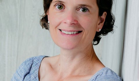 Eine Frau in niedrigem mittleren Alter mit eher kurzen braunen Haaren und einem schmalen und freundlichem Lächeln.