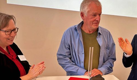 Ein Mann etwas höheren mittleren Alters in einem blauen Hemd mit kurzer weißer Halbglatze steht vor seiner Auszeichnung: Der Goldenen Stimmgabel