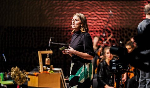 Die Frau im jungen mittleren Alter moderiert vorm Orchester stehend. Die aus vielen unregelmäßgen Akustik-Panelen bestehende Elbphilharmonie-Rückwand ist rost-rot angeschienen.