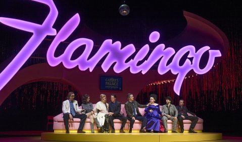 Wie in einer Star-Show aus den 70ern sitzt eine Gruppe selbstbewusster Menschen in bunter Abendrobe auf einem Sofa unter einem riesigen lila Neon-Schriftzug: "Flamingo"