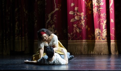 Carmen in traditionellem Kostüm hält Don José mit ihrem Körper spielend zu Boden. Im Hintergrund ein blumenreich bestickter Vorhang.