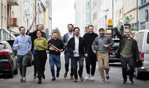 Auf einer Straße kommen uns 9 nebeneinanderer laufende Musiker:innen beziehungsweise Künstler:innen in Alltagskleidung entgegen. Sie sind mit E-Gitarre, Trompete, Querflöte, Mallet-Schlägel und Tambourin bewaffnet