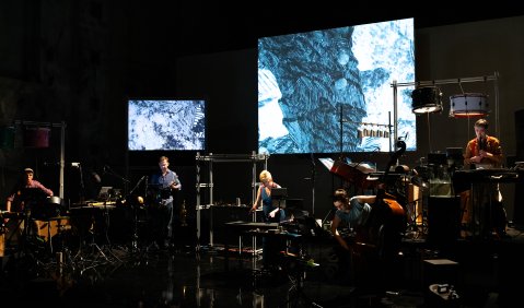 Eine dunkle Bühne. Allerlei Percussion-Instrumente, Trompeten, Theremin, Kontrabass und Flöten. Natürlich mit den entsprechenden Musiker*innen. Im Hintergrund zwei Projektionen bzw. Bildschirme die etwas zeigen, dass einer in blau gezeichneten Felssäule ähnelt.