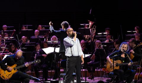 Ein Orchester in dunkel-violettem Licht. Davor am Mikrofon Sebastian Horn mit Brille, Glatze und vollem Bart in weißem Hemd mit Hosenträgern. Links und rechts flankiert von Musikern aus seiner Band.