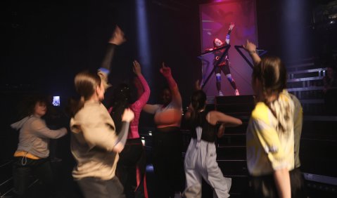 Eine Gruppe junger Menschen tanzt in unterschiedlichesten Alltagsklamotten. Vor ihr (von der Kamera aus gehesen dahinter) auf einem kleinen Podest tanzt eine Frau in schwarz mit Ketten an das Podest gezwungen.