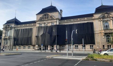Ein breites schlossähnliches Gebäude um 1900. Die Mitte der Fassade ist nicht zu sehen, sie ist von einer schwarzen Folie überspannt.