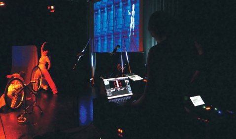 Dunkles Bühnengeschehen: Erleuchtet ist ein digitales Bühnentechnik Instrumente, eine Projektion an der Bühnenrückwand die viel blaue Fläche und einen Menschen zeigt. Vorne auf der Bühne in Rot sieht man ein Fahrrad und zwei Menschen daneben.