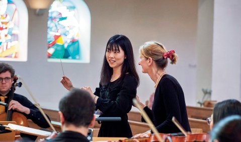 Orchesterleitungsunterricht in einer Kirche: Eine Frau gibt einer jüngeren Frau mit Taktstock Feedback für ihr Dirigat/ihre Probe mit einem Orchester.