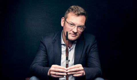 Ein Mann in Sakko, Jeans und Joggingschuhen sitzt in einem Sessel vorgelehnt und hält einen Kontrabassbogen zwischen seinen Händen vor sich.