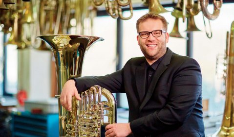 Ein Mann niedrigeren mittleren Alters sitzt mit schwarzem Anzug und eine Tuba neben sich stehend inmitten vieler Blechblasinstrumente in einer entsprechenden Musikinstrumentenwerkstatt.
