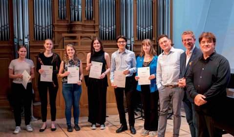 Ein Gruppe von jungen Erwachsenen steht in einem Halbkreis vor einer Orgel. Die meisten halten ein Dokument in den Händen.