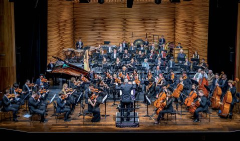 Ein großes Sinfonieorchester mit Flügel (im Orchester) in einem Konzertsaal mit Holz-Akustik-Rückwand als Spielzimer.