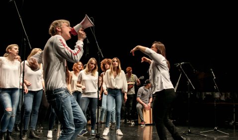 Ein Chorauftritt. Neben dem Chor und der Chorleiterin singt/ruft auch ein junger Mann in ein Megaphon und am anderen Ende der Bühne spielt jemand eine Cajon.