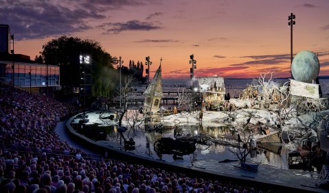 Eine große Open-Air-Bühne mit einer untergegangenen alten Kleinstadt als Bühnenbild. Im Hintergrund die reale Abendsonne mit See.