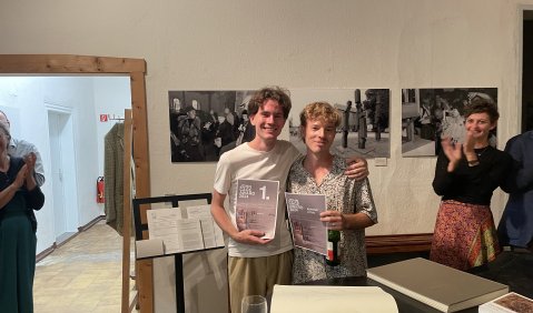 Zwei junge Musiker mit dunklen und blonden Locken stehen in locker-sommerlicher Kleidung mit ihren Urkunden in der Hand zusammen.