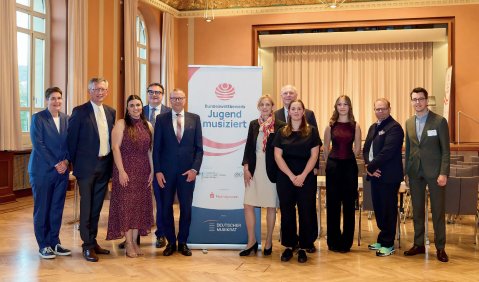 Eine Gruppe Erwachsener und einzelner jugendlicher in schicker Abendrobe in einem Schlosssaal mit Parkett und Jugend-musiziert Aufsteller.