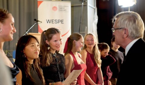 Ein Mann im höheren mittleren Alter steht mehreren jungen Jugend musiziert-Teilnehmerinnen gegenüber. Im Hintergrund ein Transparent: "WESPE"