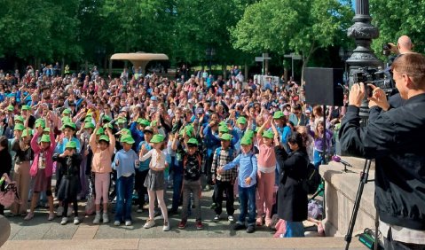 Eine große Menschenmenge unter blauem Himmel. Im Hintergrund hinter Baumkronen gläserne Wolkenkratzer.