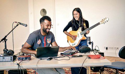 Eine E-Gitarristin und ein Producer am Laptop und Synthesizern in einem Raum.